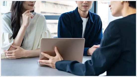 ビジネス風の服装の3人がテーブル越しにノートパソコンを見ながら話し合っている打ち合わせの様子